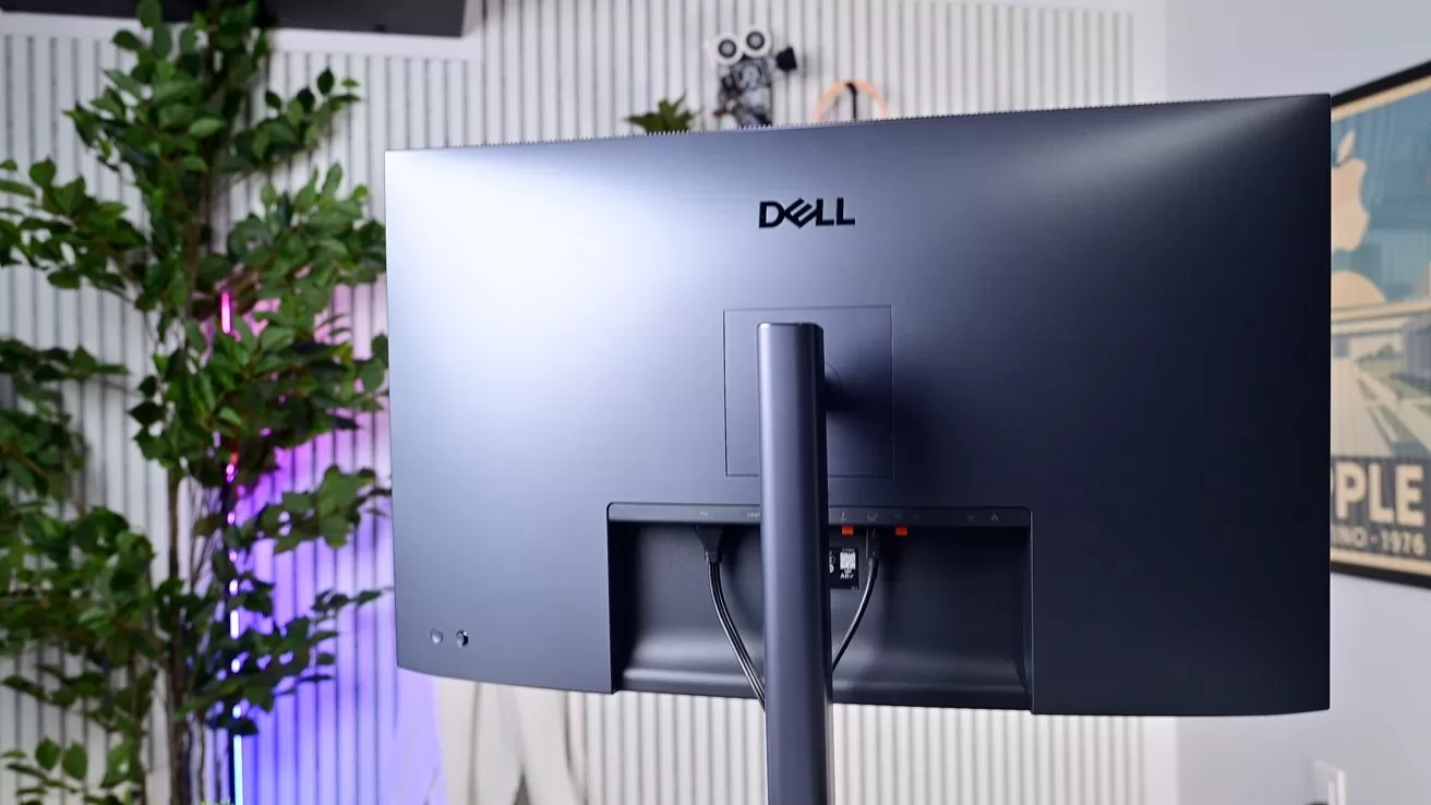 Back view of a curved Dell computer monitor on a stand, showing cable area, with a potted plant and wall art in a modern, softly lit office setting