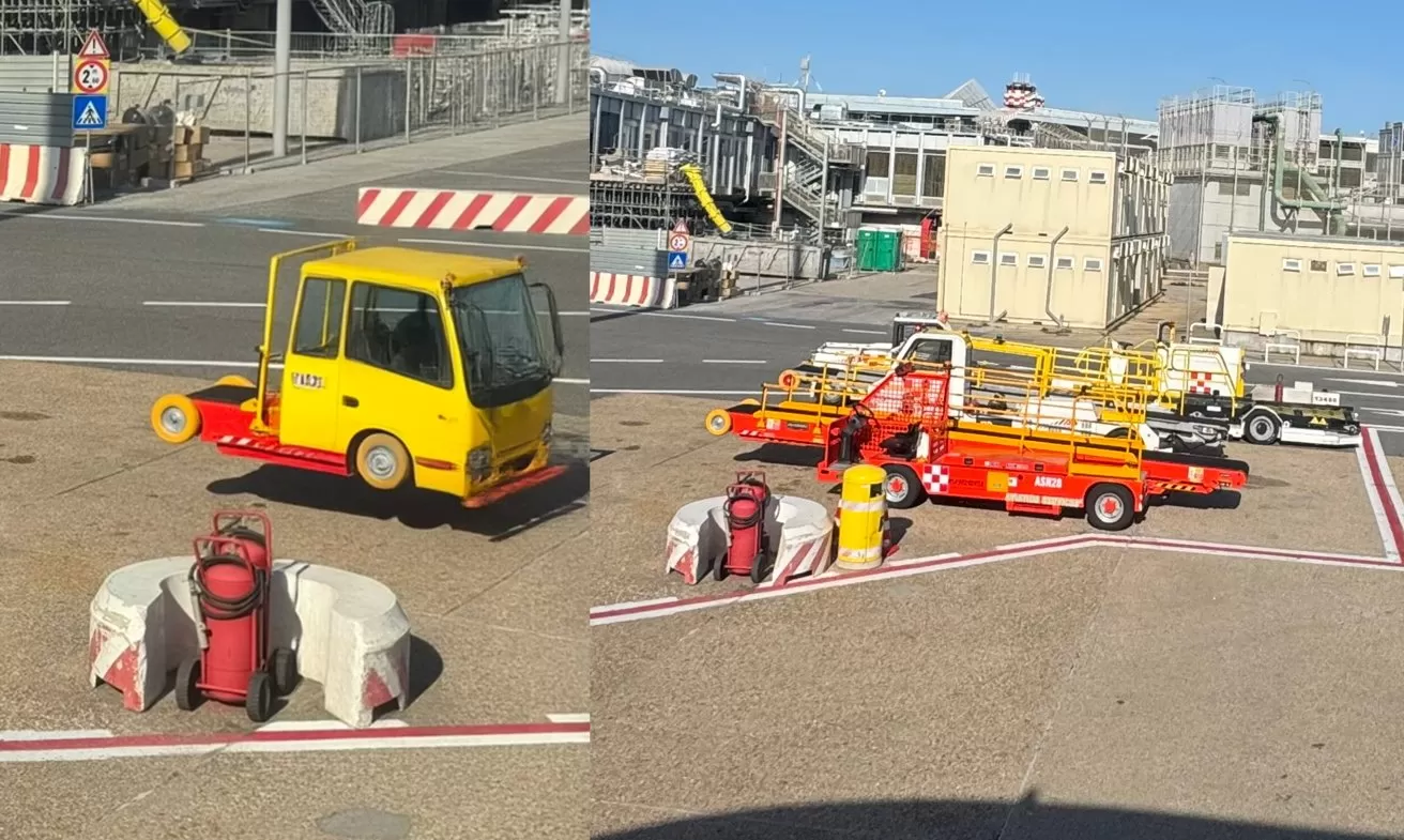 Airport service vehicles on a tarmac, including small yellow and red tow trucks and equipment carts, with safety barriers, gas cylinders, and industrial buildings in the sunny background