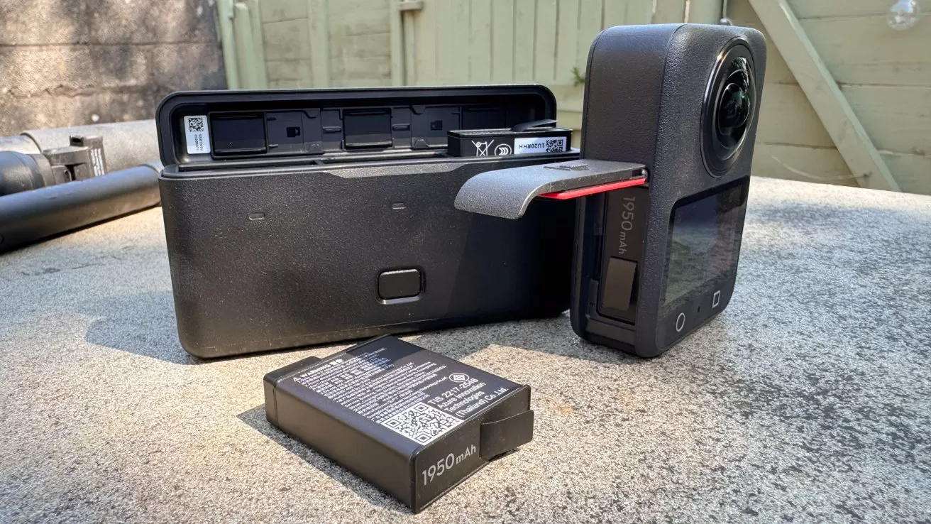 360 action camera with side door open showing battery slot, a separate rectangular battery charger behind it, and another spare 1950 mAh battery lying on a concrete surface outdoors
