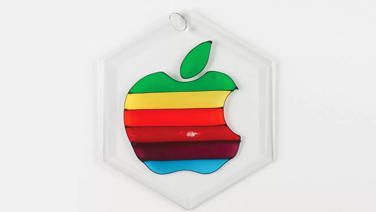 Colorful bitten apple logo with horizontal rainbow stripes, including green leaf, placed on a clear hexagonal ornament against a plain white background.