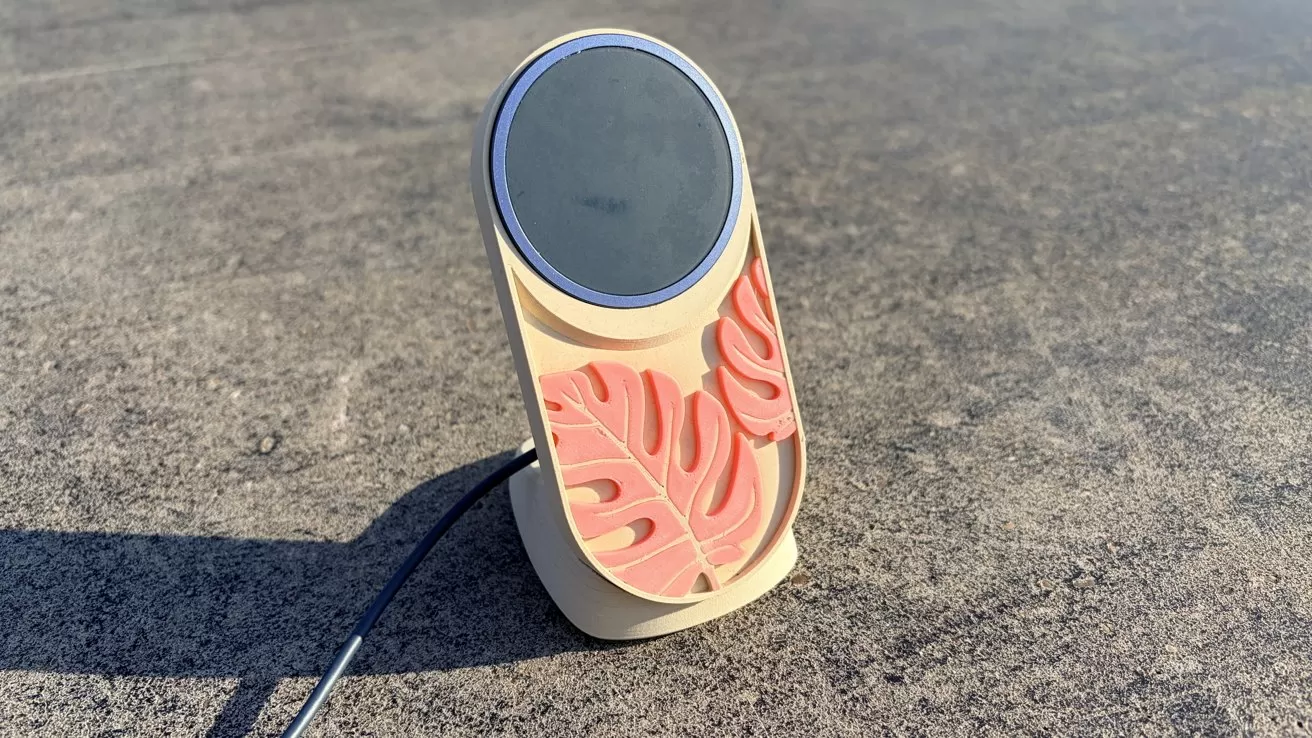 Beige upright wireless phone charger with a dark circular charging pad and decorative coral-colored monstera leaf shapes, sitting on a rough gray concrete surface in sunlight