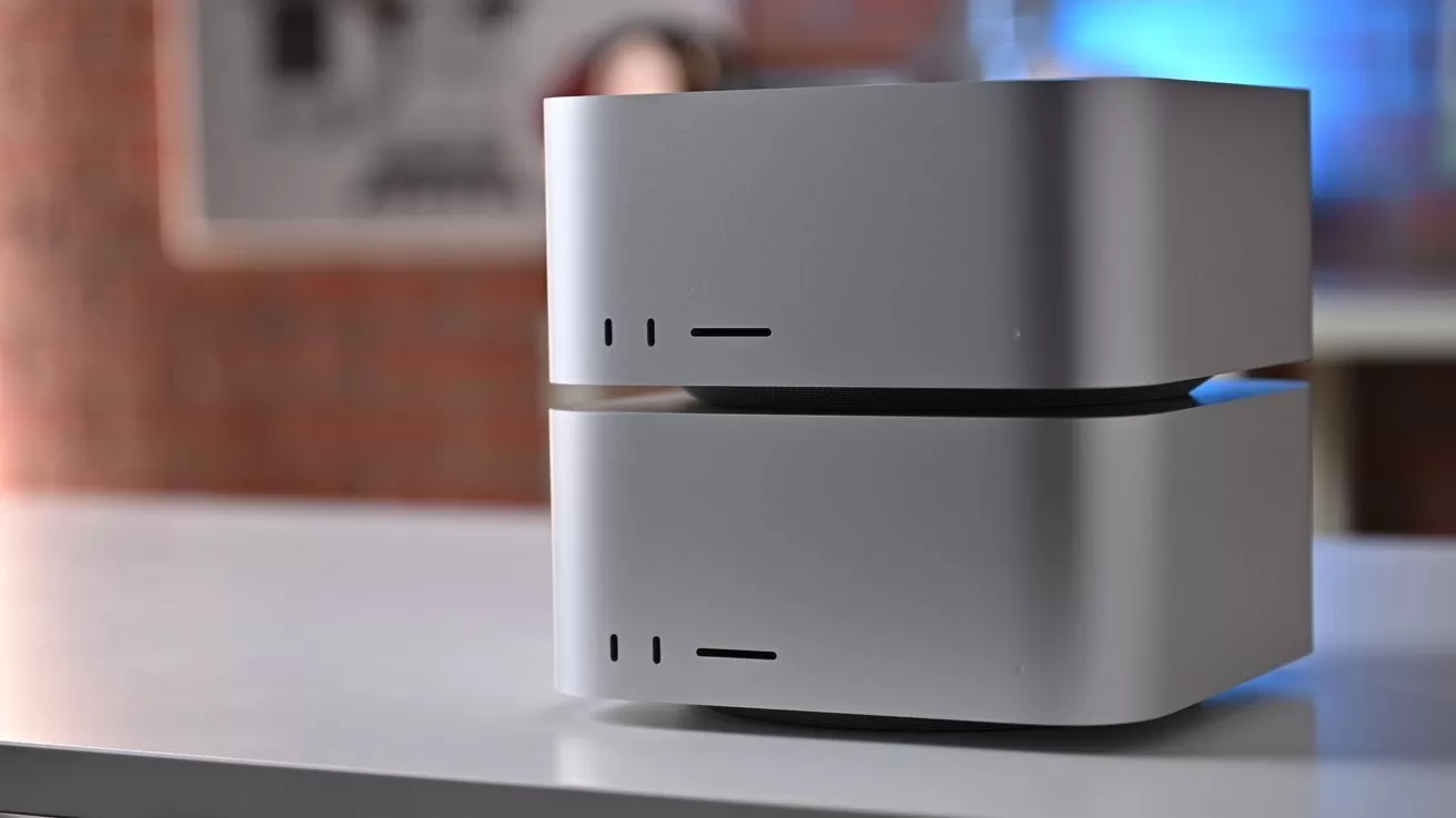 Two stacked silver Mac Studio desktop computers on a white table, showing front ports and vents, with a softly blurred brick and blue-toned background