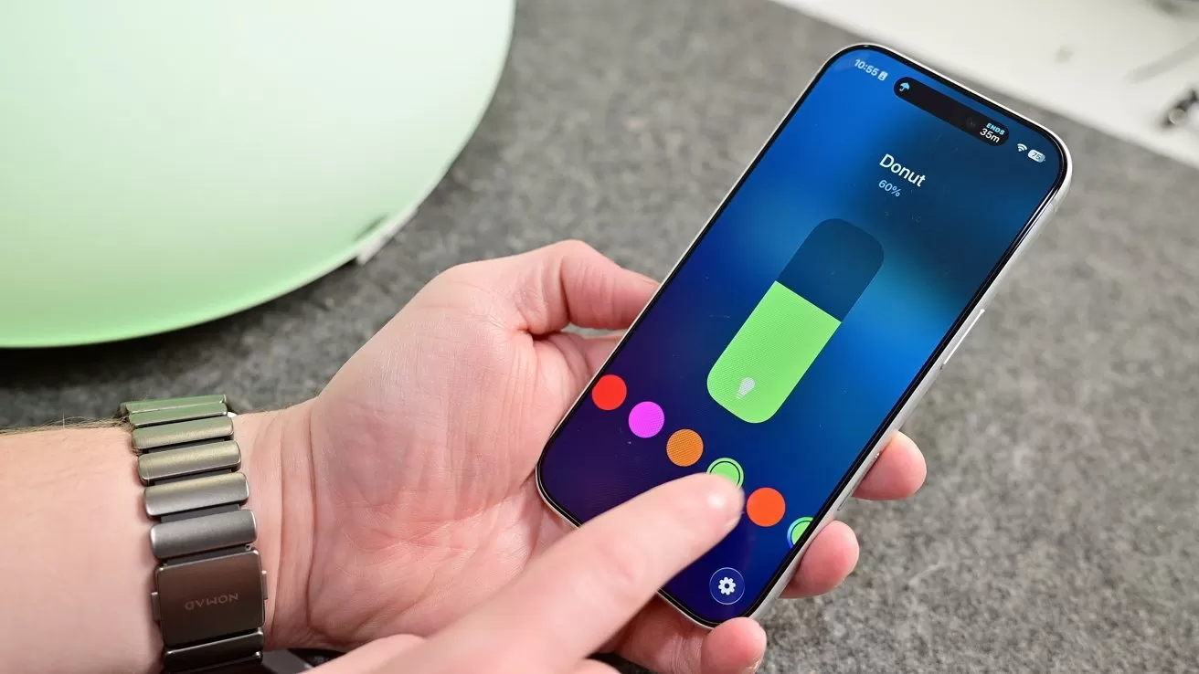 Person holding a smartphone, adjusting a colorful on-screen control slider with one hand, while the other hand supports the phone on a gray surface with a green object blurred in the background