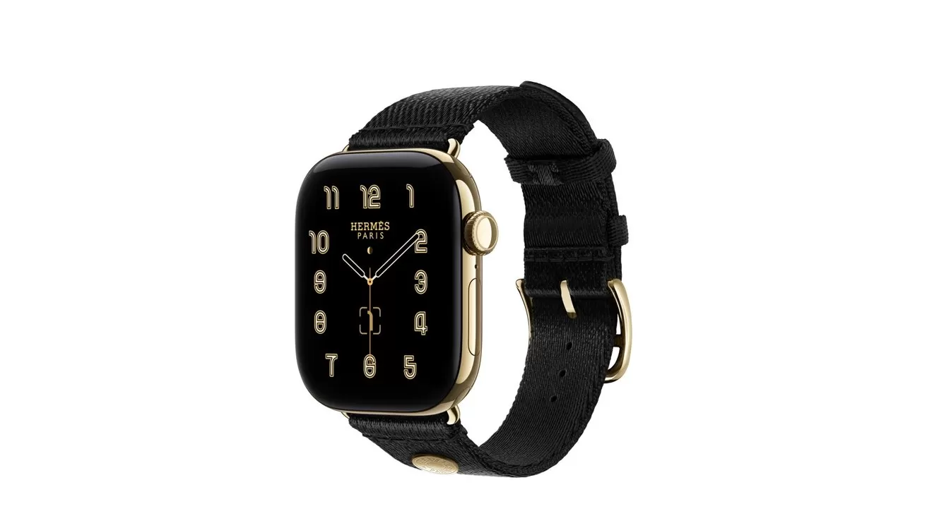 Gold smartwatch with black Hermes face, large cream Arabic numerals, and black fabric band, shown at an angle on a white background