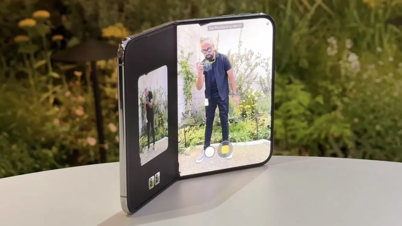 Foldable smartphone partially open on a table, using its large inner screen as a camera viewfinder showing a man posing outdoors, with greenery background and smaller preview display on the left