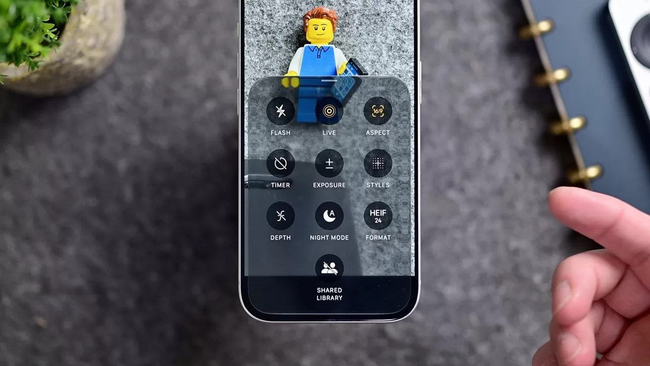 Smartphone displaying camera settings over a Lego minifigure on gray surface, with a hand reaching from the right and a small potted plant and notebook partially visible nearby