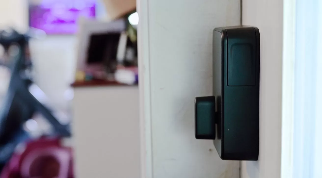 Close-up of a black smart door lock mounted on a white door, with a blurred indoor room and furniture visible in the background
