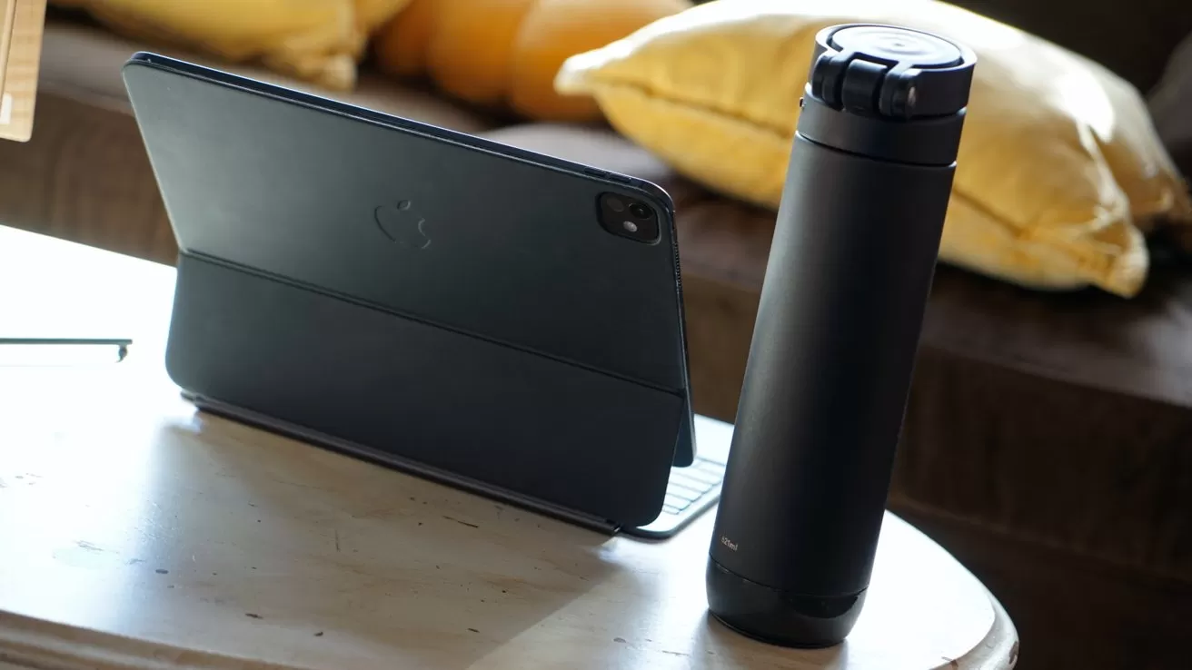 Tablet in a black case on a table beside a tall, black travel mug, with yellow cushions in the background.