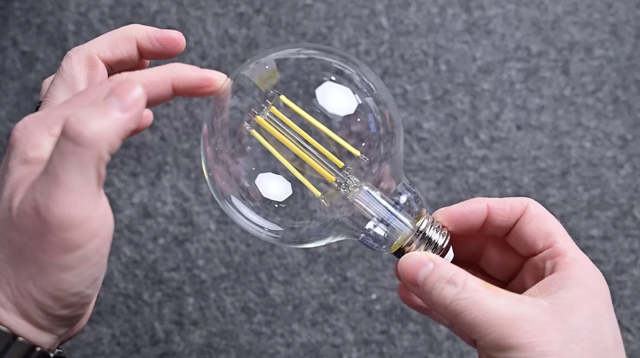 Two hands holding a large clear LED filament light bulb against a gray textured background, with fingers gently touching the glass near the top of the round bulb